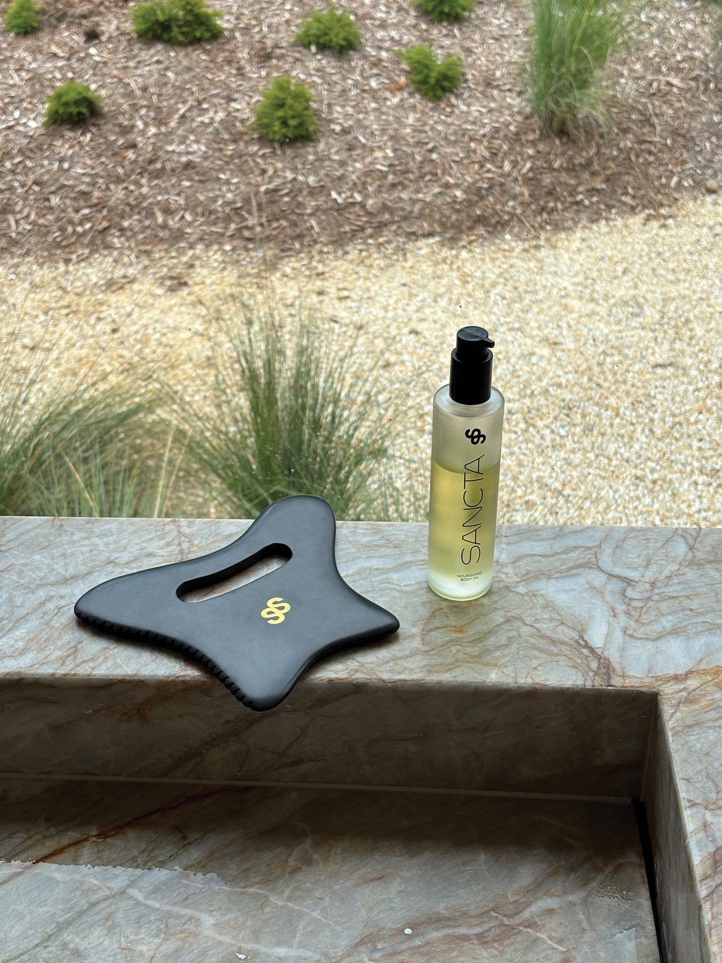 Sancta Solis Duo (Bian stone body gua sha tool and nourishing body oil) pictured in an outdoor bathroom on a marble sink surrounded by foliage.