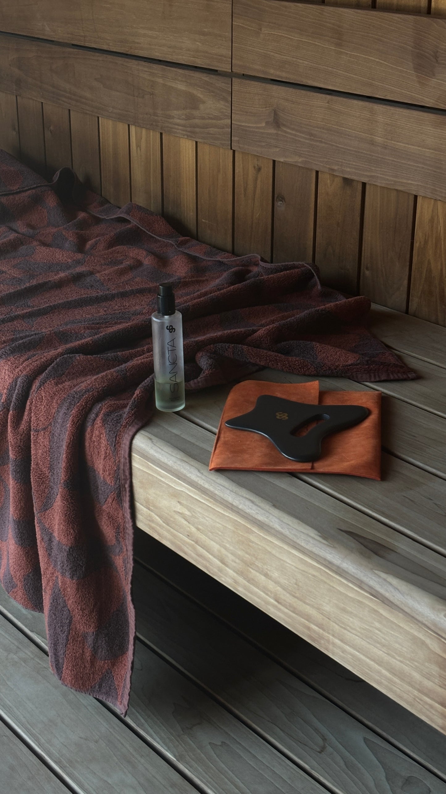Sancta Solis Bian stone Body Gua Sha tool  sitting with the Nourishing Body oil sitting next to a brown patterned towel in a cosy wooden Sauna.