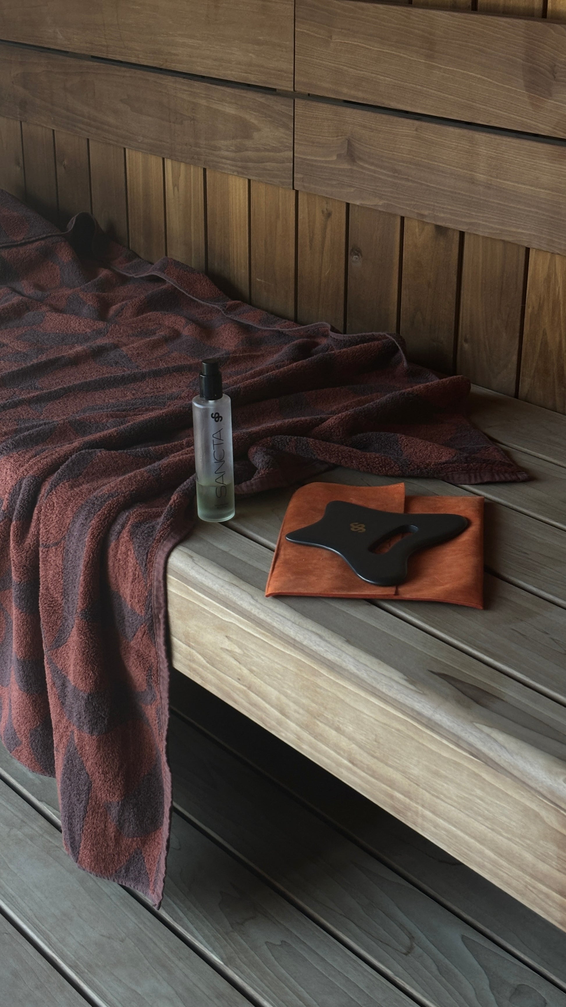 Sancta Solis Bian stone Body Gua Sha tool  sitting with the Nourishing Body oil sitting next to a brown patterned towel in a cosy wooden Sauna.