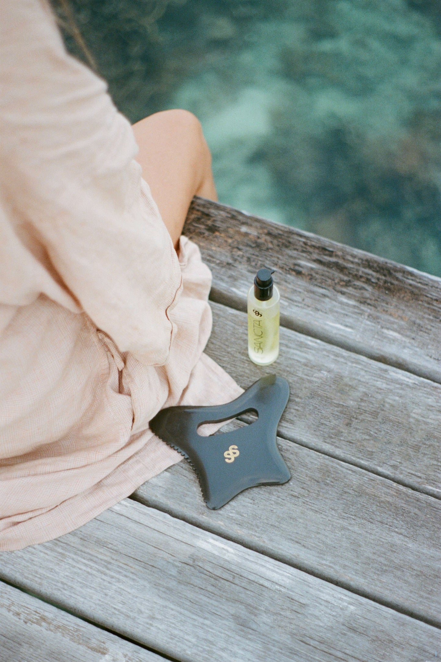 A woman sitting on a jetty by the ocean. Next to her is the Sancta Solis Nourishing Body Oil bottle and a Sancta Solis Bian Stone Body Gua Sha Tool