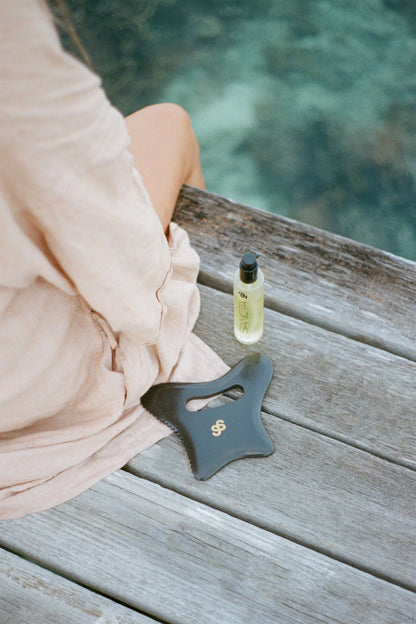 A woman sitting on a jetty by the ocean. Next to her is the Sancta Solis Nourishing Body Oil bottle and a Sancta Solis Bian Stone Body Gua Sha Tool
